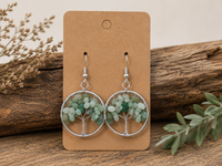 A pair of dangling tree-of-life earrings with light green and white beaded foliage inside silver circular frames, displayed on a brown kraft card against a rustic wood background with dried flowers and greenery.
