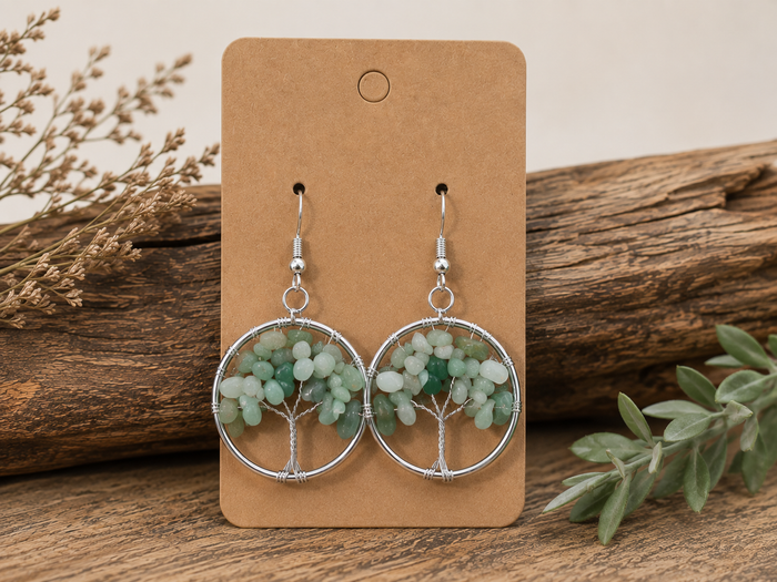 A pair of dangling tree-of-life earrings with light green and white beaded foliage inside silver circular frames, displayed on a brown kraft card against a rustic wood background with dried flowers and greenery.