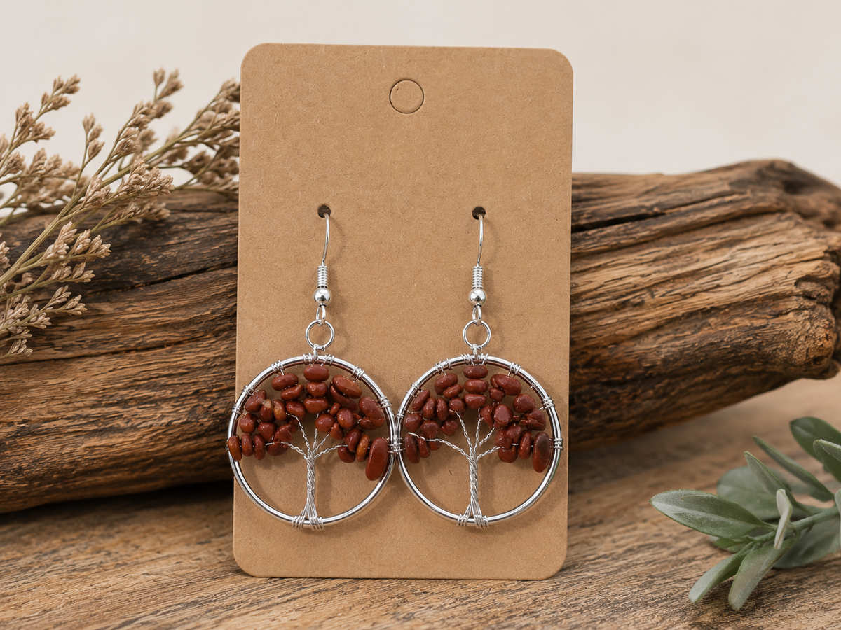 A pair of dangling earrings displayed on a tan card, set against a rustic wood background with dried flowers and green leaves. The earrings feature silver-tone circular wire frames with a tree design made from reddish-brown seed beads.