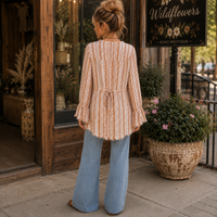 A woman stands on a sidewalk in front of a boutique, viewed from behind. She is wearing a long-sleeve, tie-waist, cream and peach patterned top with flared bell sleeves, paired with light blue wide-leg jeans.
