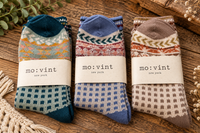 Three pairs of folded knit socks with patterned tops and checkerboard lower sections, each wrapped with a paper band reading 'mo:vint new york,' arranged on a rustic wooden surface with decorative dried flowers and greenery.