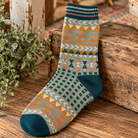 A single knit sock with a multicolor geometric and tribal pattern, resting on a rustic wooden surface next to a wooden block and some green foliage.