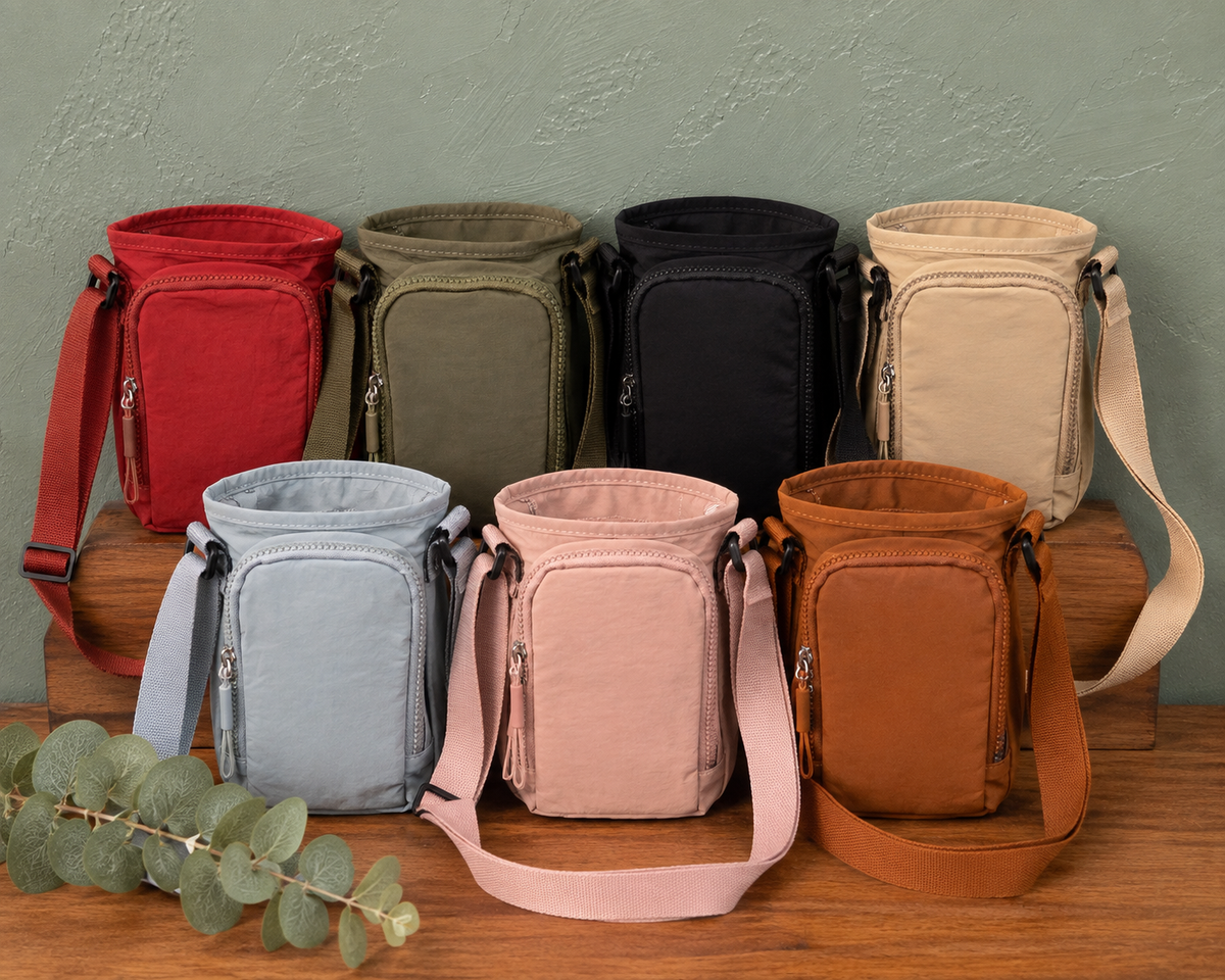 Six water bottle crossbody bags in red, olive green, black, beige, light blue, pink, and brown, arranged on wooden blocks against a textured green wall.