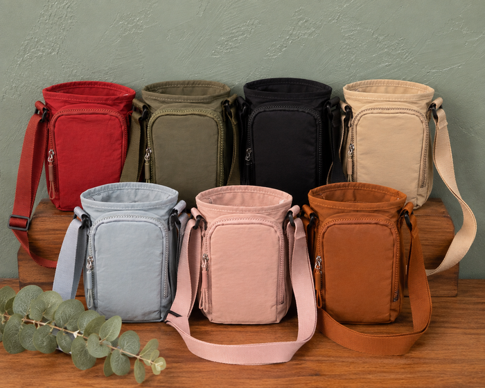 Six water bottle crossbody bags in red, olive green, black, beige, light blue, pink, and brown, arranged on wooden blocks against a textured green wall.