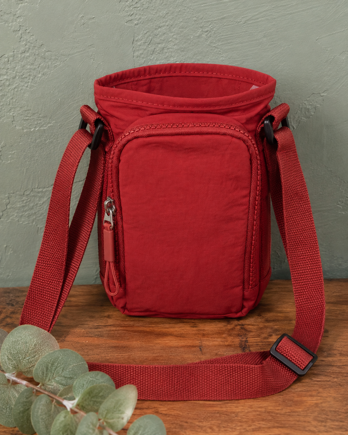 A red nylon crossbody pouch bag with a front zip pocket and adjustable shoulder strap, displayed on a wooden surface against a textured gray wall with eucalyptus leaves in the foreground.