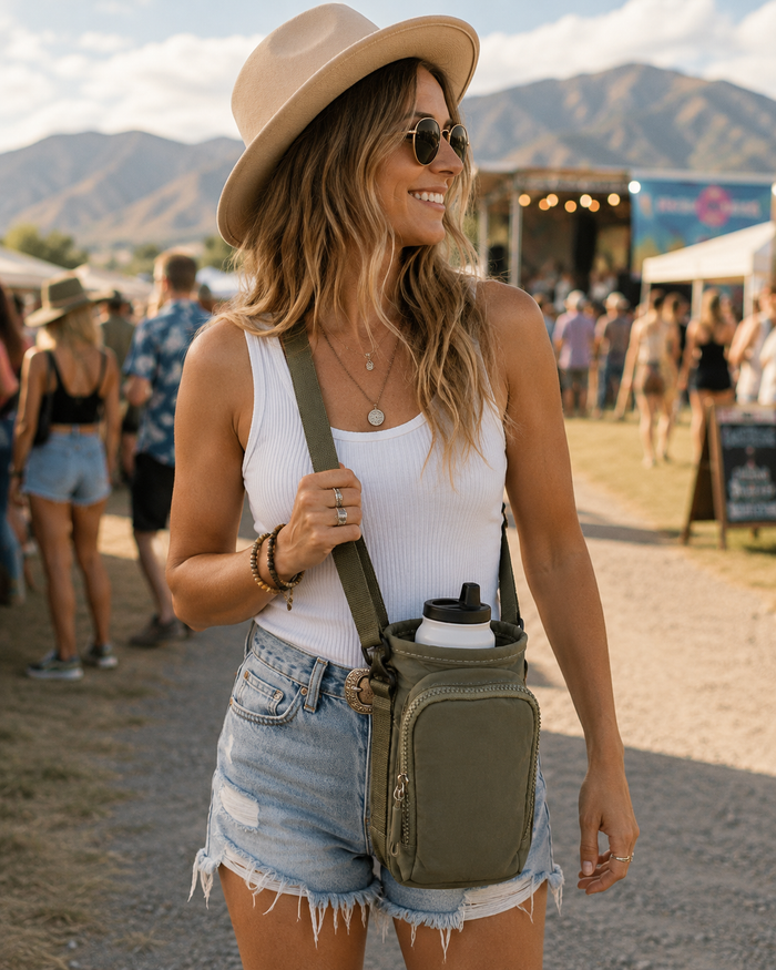 A woman at an outdoor festival wearing a beige wide-brim hat, white ribbed tank top, and denim shorts, carrying an olive green crossbody bottle bag with a water bottle.