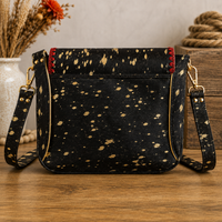 Black crossbody handbag with a gold speckled pattern, gold trim, and red stitching accents, displayed on a wooden surface with rustic decorative props in the background.