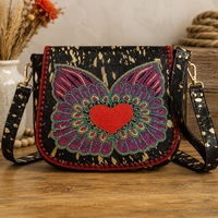 A black crossbody bag with a colorful beaded butterfly and red heart design on the front, displayed on a wooden surface with rustic decorative props in the background.