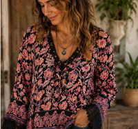 A woman wearing a black long-sleeve bohemian-style blouse with a vibrant pink, orange, and blue floral pattern, lace trim, and a V-neckline, set against a rustic indoor background with plants.