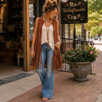 A woman walking on a brick sidewalk in front of a boutique, wearing a rust-brown fringed sleeveless vest over a cream lace camisole, blue distressed flare jeans, and tan sandals. A sign for Wildflowers Boho Boutique is visible in the background.