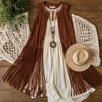 A bohemian-style outfit flat lay featuring a sleeveless rust-brown fringed vest layered over an off-white dress, styled with a cream crochet shawl, a beige wide-brim hat, and green foliage on a rustic wooden background.