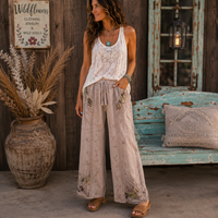 A woman stands outdoors in front of a rustic wooden wall and bench, wearing a white sleeveless lace top and loose beige wide-leg pants with floral embroidery and a drawstring waist. She is wearing tan sandals and layered necklaces.