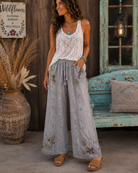 A woman stands in a rustic outdoor setting wearing a white crochet tank top and light gray wide-leg pants with floral embroidery. She is posed in front of a weathered wooden wall, a turquoise bench, and a large decorative vase.