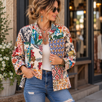 A woman wearing a colorful bohemian-style patchwork jacket with floral and geometric patterns, styled with a white top and blue jeans, standing in front of a storefront.