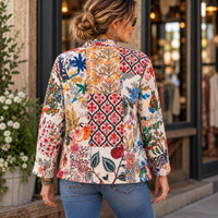 A woman viewed from behind wearing a long-sleeve patchwork quilted jacket with a vibrant multicolor floral and botanical pattern on a cream background, paired with blue jeans in front of a storefront.